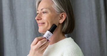 A photo of Michele, a customer of Simple Body looking to the side and smiling and holding a bottle of Simple Body Age Defense Serum.