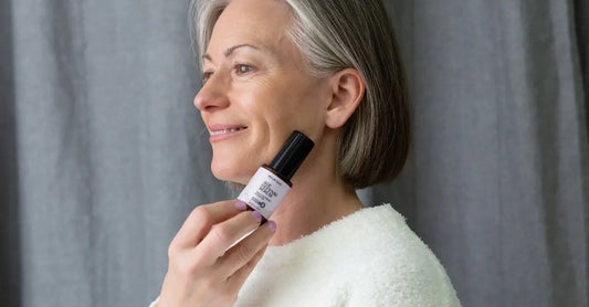 A photo of Michele, a customer of Simple Body looking to the side and smiling and holding a bottle of Simple Body Age Defense Serum.