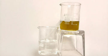 Two glass beakers one containing Vegetable Glycerin for dry skin and the other JoJoba oil for dry skin on a beige colored background.