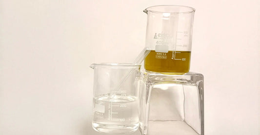 Two glass beakers one containing Vegetable Glycerin for dry skin and the other JoJoba oil for dry skin on a beige colored background.