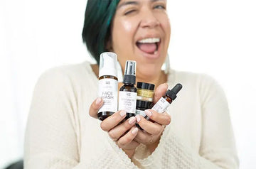 A photo of Ani, a Simple Body customer smiling big and holding up a Simple Body Face Wash, Simple Body Rosewater Toner, Simple Body Beauty Balm, Simple Body Face Cream and a Simple Body Nightly Face Serum all in travel sizes.