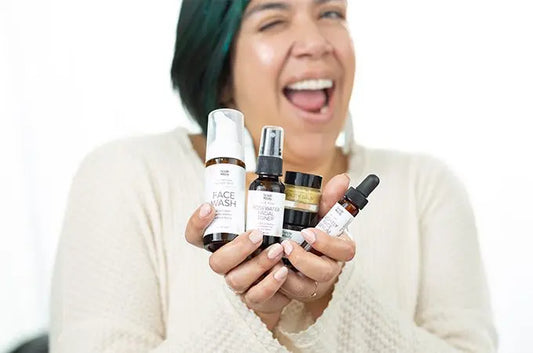 A photo of Ani, a Simple Body customer smiling big and holding up a Simple Body Face Wash, Simple Body Rosewater Toner, Simple Body Beauty Balm, Simple Body Face Cream and a Simple Body Nightly Face Serum all in travel sizes.