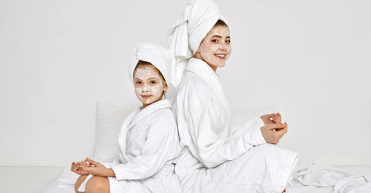 A mom and daughter sitting back to back in white robes and white Kaolin Clay masks on and towels on their heads on a white backgroudn.