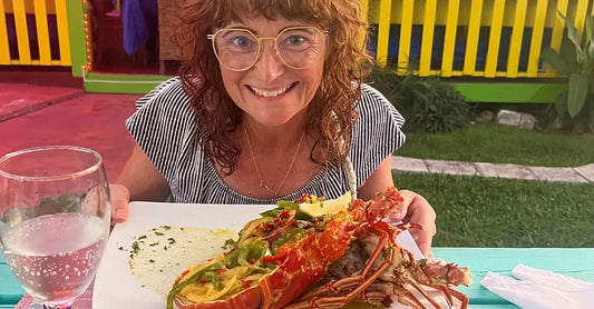 Jewels Burdick, Founder of Simple Body sits at a table in front of a lobster stuffed with fresh vegetables and a glass of water with a vibrant yellow fence in the background.
