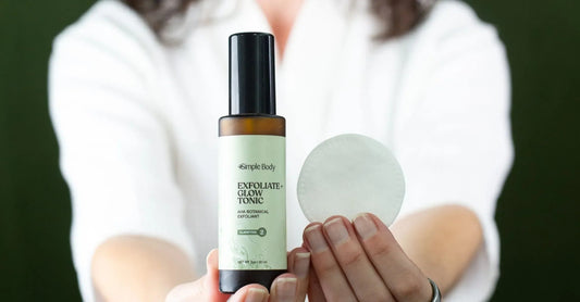 A close up of Simple Body's Exfoliate and Glow with a woman holding it and a cotton pad in the other hand in a white robe using it to treat hormonal acne on a dark green background.