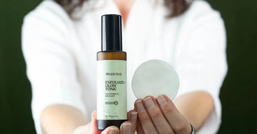 A close up of Simple Body's Exfoliate and Glow with a woman holding it and a cotton pad in the other hand in a white robe using it to treat hormonal acne on a dark green background.