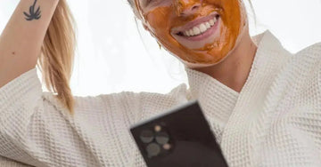 A photo of Caityln, a Simple Body Customer wearing a Pumpkin Enzyme Face Mask in a white waffle cotton robe looking at her phone and smiling.