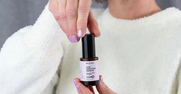 A photo of a woman wearing a white, soft looking sweater holding a bottle of Simple Body Age Defense Serum. She has purple painted nails that match the color of the label. 