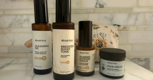 A photo of four Simple Body products sitting in front of a vintage floral cosmetic bag surrounded by marble tile including Simple Body Cleansing Oil, Simple Body Brighten and Boost Tonic, Simple Body Barrier Defense Serum, and Simple Body Face Cream