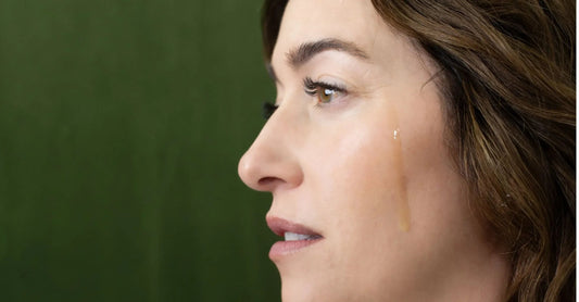 A woman around 40 years old looks to the side with a Simple Body Serum on her cheek and a dark green background.