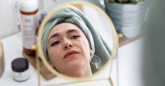 A photo of Sarah, a Simple Body customer with her hair wrapped in a towel looking in a gold mirror with Simple Body Face Wash blurred in the background.