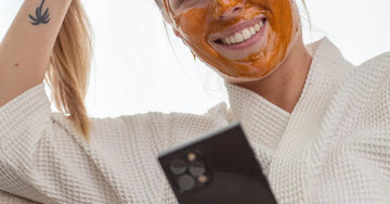 A photo of Caityln, a Simple Body Customer wearing a Pumpkin Enzyme Face Mask in a white waffle cotton robe looking at her phone and smiling.