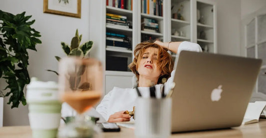 A photo of a woman with her hand on her head looking like she is stressed out sitting in front of her computer with a cup of coffee and her eyes closed.