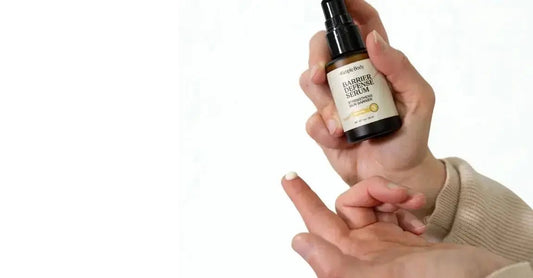 A photo of a hand holding a bottle of Simple Body Barrier Defense Serum. There's a drop of the Barrier Defense Serum on her fingertip showing off the creamy white texture.