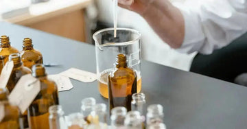 A photo of a chemist dropping oil into a beaker with some amber colored bottles on the table