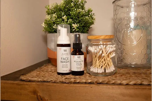 A photo of Simple Body travel size Rose Face Wash, Rosewater Facial Toner, a jar of qtips, a beautiful green plant and a candle in a glass jar sitting on a jute matt.