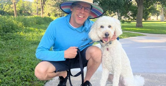 Dave kneeling next to his white poodle Bayleigh with a green park and sidewalk in the background.