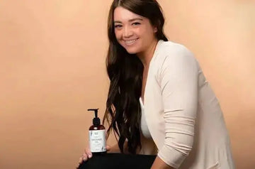 A beautiful, young woman sitting and smiling and holding a bottle of Simple Body Massage Oil with a warm beige background.