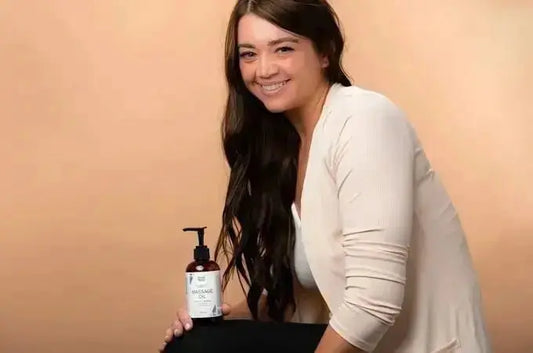 A beautiful, young woman sitting and smiling and holding a bottle of Simple Body Massage Oil with a warm beige background.