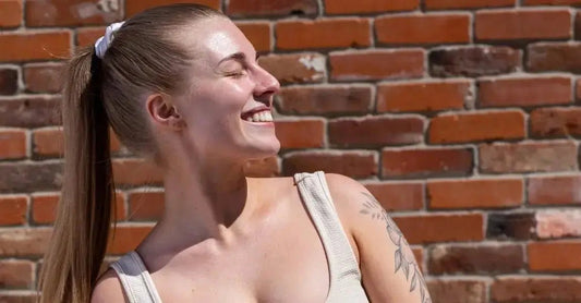 A photo of Caitlyn looking into the sunshine and smiling. She wears a ponytail for her long hair and an off white tank top and sits in front of a brick wall.