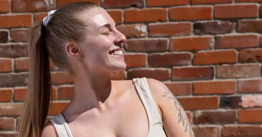 A photo of Caitlyn looking into the sunshine and smiling. She wears a ponytail for her long hair and an off white tank top and sits in front of a brick wall.