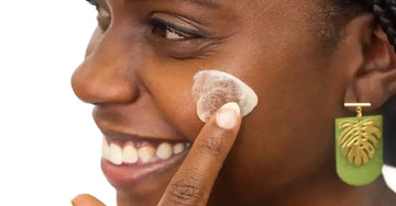 A photo of a young woman applying Simple Body Face Cream to her cheek smiling big.