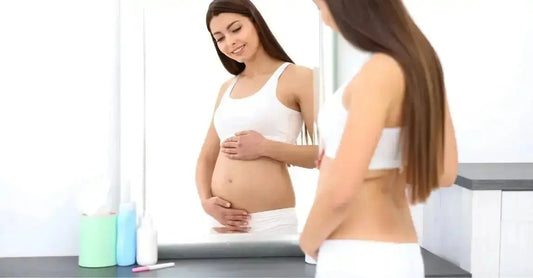 A young, pregnant woman looking in the mirror holding her belly and smiling with natural skincare products on the counter.