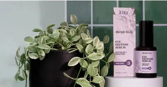 A photo of Simple Body Eye Defense Serum sitting on top of a white block with a green tile background and a healthy green plant next to it.