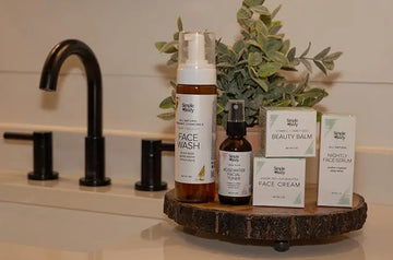 A photo of Simple Body products sitting on a wooden tray including Chamomile Foaming Face Wash, Rosewater Facial Toner, Beauty Balm, Face Cream and Nightly Face Serum with a green plant in the background and a black modern faucet.