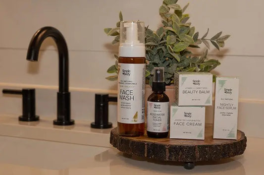 A photo of Simple Body products sitting on a wooden tray including Chamomile Foaming Face Wash, Rosewater Facial Toner, Beauty Balm, Face Cream and Nightly Face Serum with a green plant in the background and a black modern faucet.