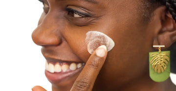 A photo of a young woman applying Simple Body Face Cream to her cheek smiling big.