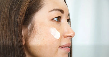A photo of a young woman looking to the side with beautiful brown hair and brown eyes. She has a swatch of Barrier Defense Serum for sensitive skin on her cheek.