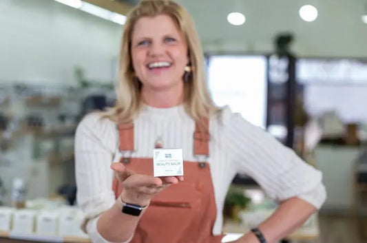 A photo of Stacie, a former employee of Simple Body smiling and wearing coral colored overalls and holding up a Simple Body Beauty Balm for age supporting skincare