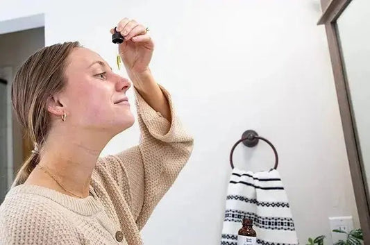 A young woman with her hair pulled back in a ponytail and a neutral colored top on looking in the mirror applying Simple Body's Pomegranate Serum