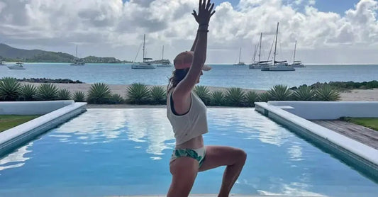 Jewels Burdick, Founder of Simple Body does a yoga pose in her bathing suit in front of a beautiful pool with the teal blue Carribean water in the background with yachts.