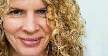 A closeup photo of Judy, a Simple Body customer, looking directly into the camer and smiling with gorgeous skin and beautiful curly blond hair.