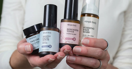 A close up image of hands holding Simple Body Face Cream, Simple Body Nightly Face Serum, Simple Body rosewater Toner and Simple Body Foaming Face Wash in chamomile with a white background.