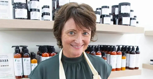 A photo of Simple Body Founder, Jewels Burdick, standing in front of her product line including Simple Body Face Cream, Simple Body Bamboo Face Polish, Simple Body Body Butter, Simple Body Lotion in 8oz and 4oz. 