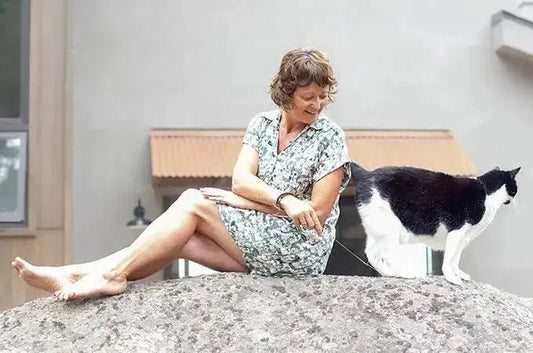 A photo of Jewels Burdick, Founder of Simple Body, sitting on a large rock playing with her tuxedo cat, Alex.