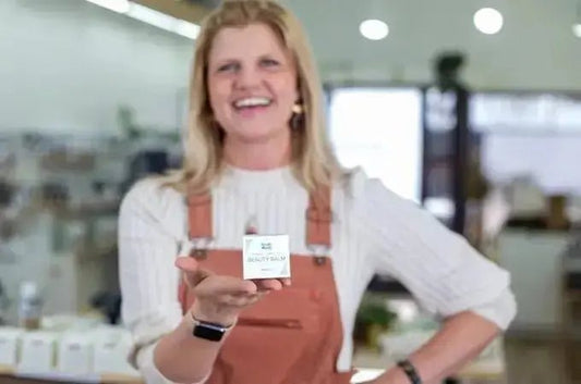 A photo of Stacie, a former employee of Simple Body smiling and wearing coral colored overalls and holding up a Simple Body Beauty Balm for age supporting skincare