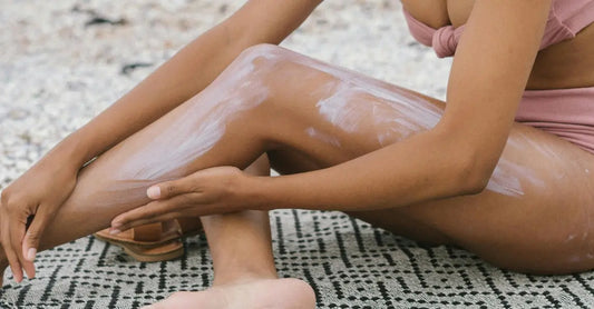 A closeup photo of a woman sitting at the beach on a patterned towel applying Think Sunscreen to her leg.