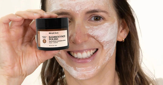 A photo of a young woman holding a Simple Body Bamboo Face Polish in Rose + Orange with some painted on her face that has a white texture and she's smiling.
