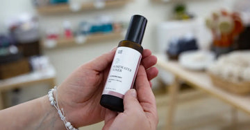 A photo of female hands holding up a bottle of Simple Body Rosewater Facial Toner with a blurred background.