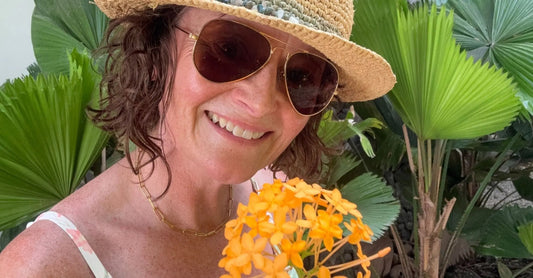 A photo of founder, Jewels Burdick of Simple Body wearing sunglasses and a straw hat holding a tropical flower standing in front of palm leaves.