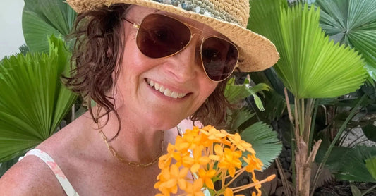 A photo of founder, Jewels Burdick of Simple Body wearing sunglasses and a straw hat holding a tropical flower standing in front of palm leaves.