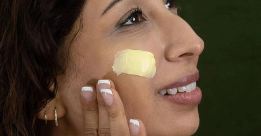 A close up of a woman's cheek with a swipe of yellow cream, known as Simple Body Beauty Balm with a french manicure smiling.