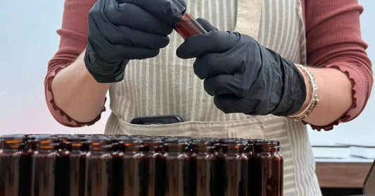 A close up photo of a woman in a beige striped apron and rust colored shirt with black gloves on holding a small amber colored bottle and filling it with Simple Body Eye Defense Serum.