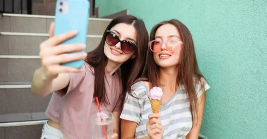 Two young women from Gen Z are taking a selfie while sitting on concrete stairs in front of a teal wall holding a coffee and an ice cream cone