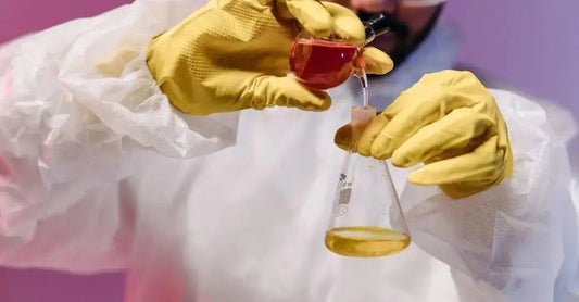 A photo of a scientist pouring a liquid from one beaker to another and wearing yellow gloves and a plastic apron.