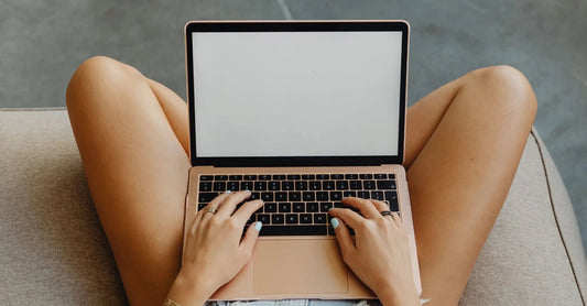 A photo of a woman from the top down sitting cross legged on a beige couch in front of a laptop with a blank screen.